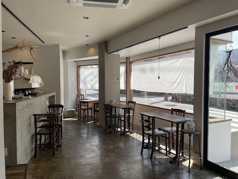 A bright, minimalist cafe interior. Concrete floors and counter pair with dark wooden tables and chairs under natural light from large windows. Dried branches offer a rustic touch. An espresso machine sits ready behind the counter, hinting at fresh brews in this calm, modern space.