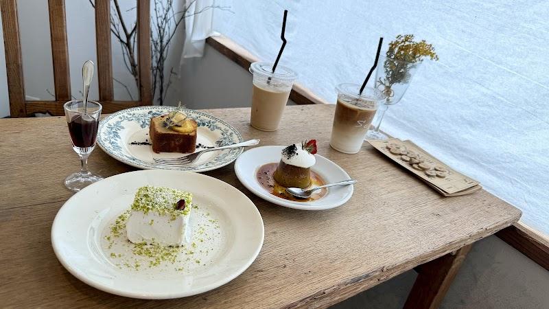 A cozy cafe table by a bright window, showcasing a rustic wooden surface. It features two iced lattes, a square pistachio-dusted cake, a caramel flan with whipped cream and strawberry, and a lemon loaf slice. A small glass of dark liquid and dried flowers complete this charming dessert spread.