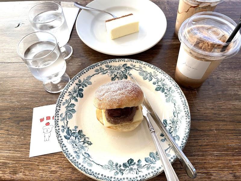 A cozy cafe table spread with sweet treats. A fluffy bun filled with red bean paste and butter, dusted with sugar, rests on a vintage-style blue floral plate. Two iced coffees/lattes, a slice of cheesecake, and water are also visible on the rustic wooden surface.