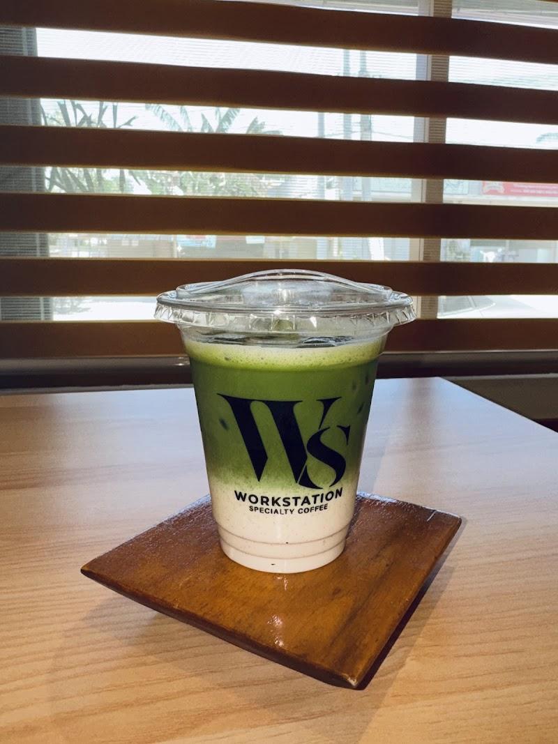 An iced matcha latte with vibrant green and white layers, from "Workstation Specialty Coffee," rests on a dark wood coaster on a light table. Bright sunlight filters through horizontal window blinds in the background, creating a peaceful cafe atmosphere.