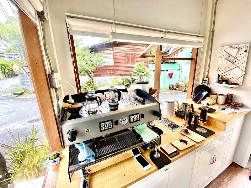 Bright, inviting cafe interior featuring a prominent espresso machine and a pour-over coffee station on a light wood counter. White cabinets below. A large window offers a view of lush greenery and rustic buildings outside, creating a relaxed atmosphere perfect for enjoying fresh brews.