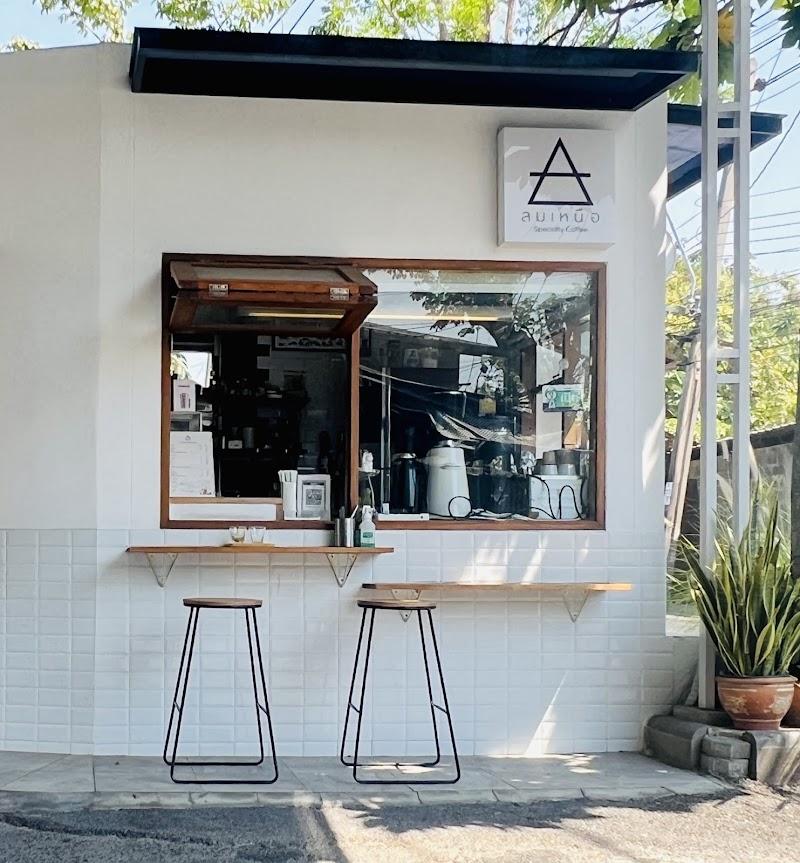 A bright, modern cafe facade with white tiled walls. A large window, partially open for service, displays coffee equipment inside. Two sleek wooden stools and a wooden counter invite outdoor patrons. A minimalist geometric logo and Thai text above suggest a specialty coffee spot with a clean, inviting atmosphere.