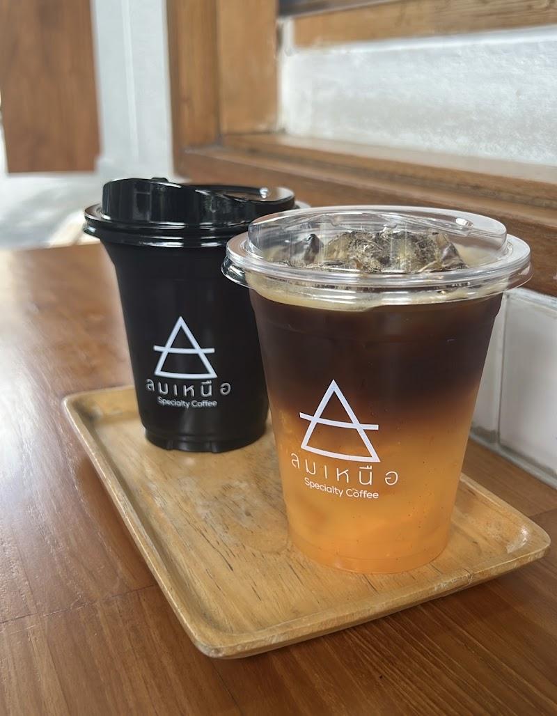 Two iced specialty coffees from 'ลมเหนือ Specialty Coffee' on a wooden tray. One is in a black cup, the other is a striking layered drink with a vibrant orange base and dark coffee on top, served in a clear cup with ice. The setting features a warm wooden table, suggesting a cozy cafe atmosphere.